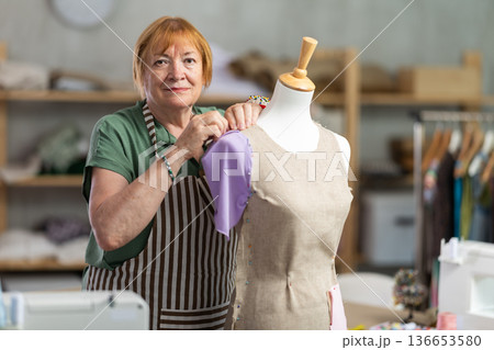 Mature female tailor sews new dress on a mannequin in sewing studio Mature female tailor sews new dress on a mannequin in sewing studio 136653580