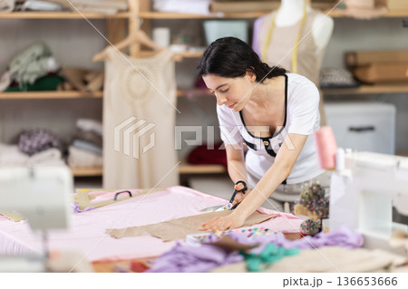 Female seamstress cutting fabric along patterns on worktable 136653666