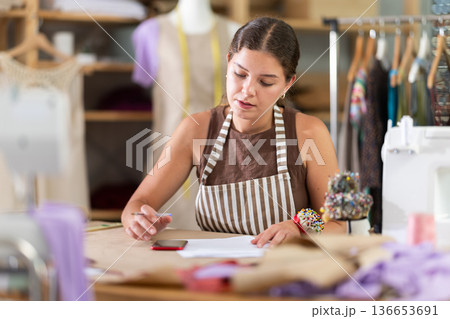Young seamstress making calculations on paper standing near table Young seamstress making calculations on paper standing near table 136653691