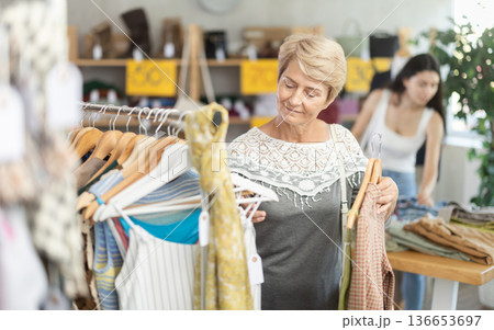 Senior woman customer elect something summer clothes in store. Female client choosing something in background Senior woman customer elect something summer clothes in store. Female client choosing something in background 136653697