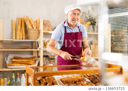 Senior man seller puts croissant in plastic disposable bag for buyer, packs goods. Senior man seller puts croissant in plastic disposable bag for buyer, packs goods. 136653725