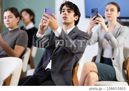 Positive young guy student sitting and listening lecture, taking photo or QR with mobil or tablet in lecture hall 136653803