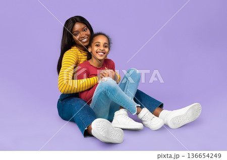 A mother and her daughter are sitting together on the floor against a purple background. They are smiling and enjoying their time together, showing affection and happiness. A mother and her daughter are sitting together on the floor against a purple background. They are smiling and enjoying their time together, showing affection and happiness. 136654249