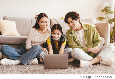 Smiling young asian female, male and teen girl sitting on floor and watching video, searching, browsing on laptop in living room interior. Video call together, modern device at home and new normal Smiling young asian female, male and teen girl sitting on floor and watching video, searching, browsing on laptop in living room interior. Video call together, modern device at home and new normal 136654399