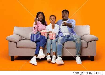 Three people sit on a couch in a bright room. They hold popcorn and look surprised. The adults are seated beside a child. They appear excited or startled. 136654400