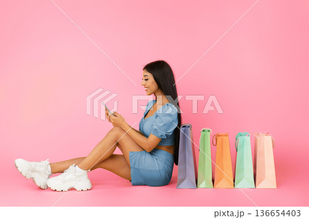 Shopping App. Laughing young woman using smartphone, sitting on floor with shopper bags, choosing and ordering clothes and presents isolated over yellow background, profile side view, free copy space 136654403