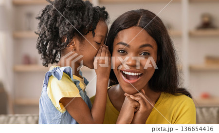 A young girl whispers to her mother while they sit in a bright living room. Both are smiling and enjoying their time together. The setting is warm and welcoming, with soft light. A young girl whispers to her mother while they sit in a bright living room. Both are smiling and enjoying their time together. The setting is warm and welcoming, with soft light. 136654416
