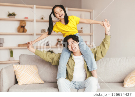 Cheerful young asian man holds on his shoulders teenager girl, sit on sofa in living room interior. Dad and daughter have fun, relationship, time together and happy family, joy at COVID-19 lockdown 136654427