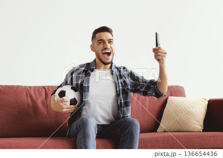 A man sits on a red couch with a soccer ball in one hand and a phone in the other. He looks excited and joyful while following a sports event on the phone. 136654436