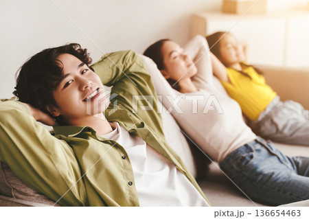 Happy youngsters japanese girl, millennial mother relax, father with hand behind head look at camera on couch in minimalist room interior. Calm, peace, rest and silence at home, family enjoy free time 136654463