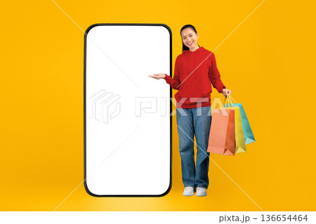 A woman stands next to a large blank phone screen, smiling and holding several colored shopping bags. The scene has a bright yellow background that adds energy to the moment. 136654464