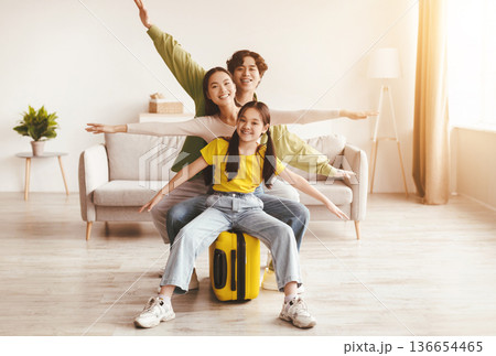 Happy excited korean young girl, mom and dad spread arms to sides like airplane, sit on yellow suitcase in living room. Preparation for travel, vacation with family, vacation after end of COVID-19 136654465