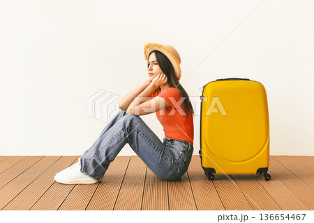 Upset female tourist sitting near suitcase and looking away at free space over light wall background. Female traveling abroad to travel on weekends getaway. Air flight journey concept Upset female tourist sitting near suitcase and looking away at free space over light wall background. Female traveling abroad to travel on weekends getaway. Air flight journey concept 136654467