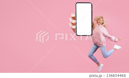 A woman is jumping with excitement while holding a big smartphone in front of her. She is smiling widely. The background is pink. This scene likely represents happiness and fun. A woman is jumping with excitement while holding a big smartphone in front of her. She is smiling widely. The background is pink. This scene likely represents happiness and fun. 136654468