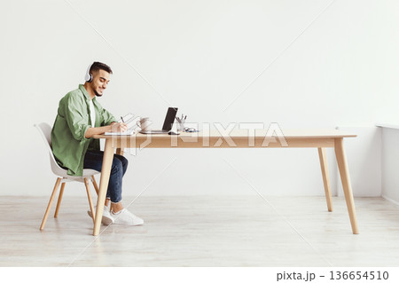 A young man sits at a wooden desk in a bright room. He wears headphones and writes in a notebook. A laptop and a cup are on the desk. The space feels open and inviting. 136654510