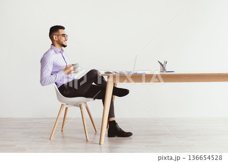 A man sits on a chair with a laptop open on a desk. He holds a large cup while looking at the screen. The room has bright walls and a minimalistic design, with stationery on the desk. 136654528