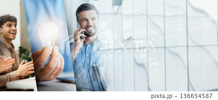 A businessman sits at his desk in a modern office. He uses a laptop while laughing and looking aside. He is engaged in online work and browsing the internet. A businessman sits at his desk in a modern office. He uses a laptop while laughing and looking aside. He is engaged in online work and browsing the internet. 136654587