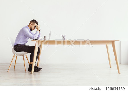A man is seated at a wooden desk, looking stressed as he holds his head in his hands. His laptop is open in front of him, and there is a cup on the table. Natural light fills the room. A man is seated at a wooden desk, looking stressed as he holds his head in his hands. His laptop is open in front of him, and there is a cup on the table. Natural light fills the room. 136654589