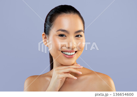 A woman with straight dark hair is smiling and resting her chin on her hand. She has clear skin and is positioned in front of a plain background. The scene shows positive energy. 136654600