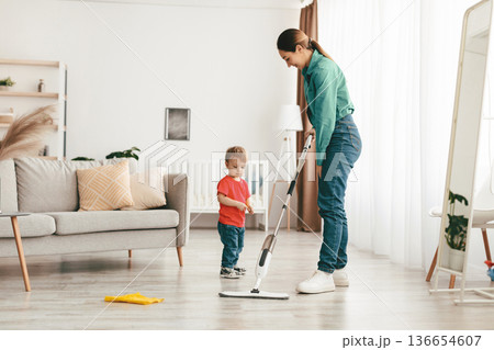 Young mother teaching toddler son cleaning at home, mopping the floor in living room interior, free space. Family housework and household concept 136654607