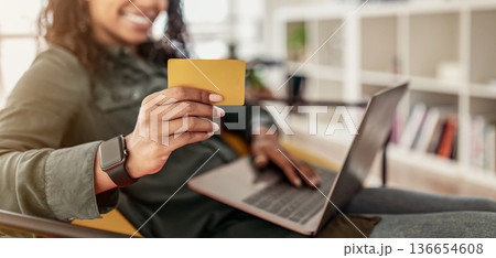 A young woman is seated in a chair, smiling as she holds a yellow credit card in one hand and types on her laptop with the other. She is inside a cozy room filled with books and plants. A young woman is seated in a chair, smiling as she holds a yellow credit card in one hand and types on her laptop with the other. She is inside a cozy room filled with books and plants. 136654608