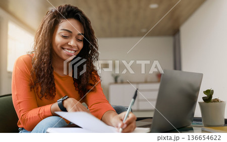 A woman writes notes on a piece of paper while seated at a table. She looks happy as she focuses on her work. A laptop is open in front of her. The room is bright and has a modern design. 136654622