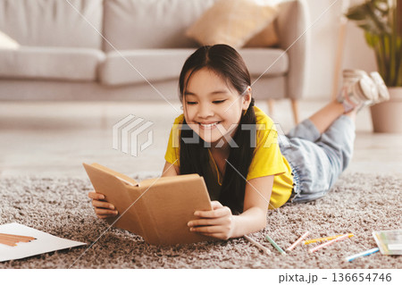 Happy teen girl lies on floor and reads book, selective focus. Young asian man and woman resting on sofa in living room interior, blurred. Relax together, fairy tales, study and family enjoy free time Happy teen girl lies on floor and reads book, selective focus. Young asian man and woman resting on sofa in living room interior, blurred. Relax together, fairy tales, study and family enjoy free time 136654746