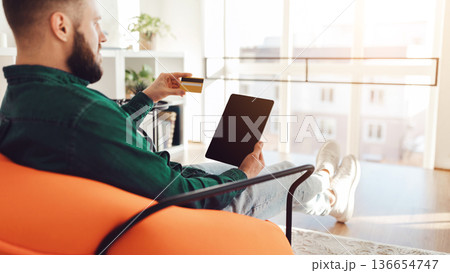 Male Buyer Shopping Online Using Digital Tablet Computer With Blank Screen Making Payment Sitting In Chair Indoors. Ecommerce Concept. Selective Focus On Display. Panorama, Side View Male Buyer Shopping Online Using Digital Tablet Computer With Blank Screen Making Payment Sitting In Chair Indoors. Ecommerce Concept. Selective Focus On Display. Panorama, Side View 136654747
