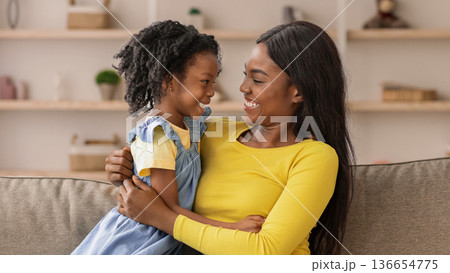 A mother sits on a couch holding her daughter while they share smiles. The living room has shelves with decorations in the background, showing a cozy home environment. A mother sits on a couch holding her daughter while they share smiles. The living room has shelves with decorations in the background, showing a cozy home environment. 136654775