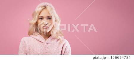 A young woman with blonde hair poses in front of a pink background. She wears a warm pink sweater. The focus is on her expression and the simple color setting to highlight her presence. 136654784