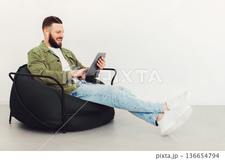Side View Of Man Using Tablet Computer And Earbuds Earphones Watching Movie Online Sitting In Chair Over White Wall Indoors On Weekend. E-Learning, Internet And Technology Concept Side View Of Man Using Tablet Computer And Earbuds Earphones Watching Movie Online Sitting In Chair Over White Wall Indoors On Weekend. E-Learning, Internet And Technology Concept 136654794