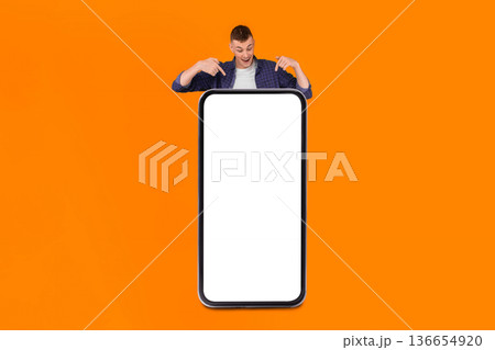 Vertical Shot Of Guy Pointing At Big Smartphone With Empty Screen Advertising New Application, Posing Standing Behind Large Cellphone On Yellow Studio Background. Mobile Technology Concept 136654920