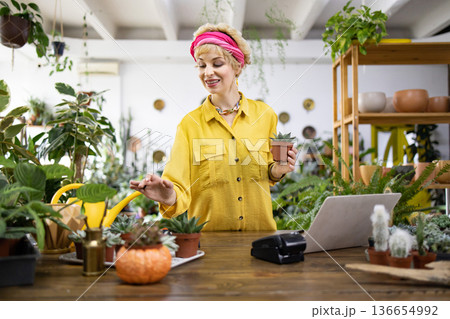 A woman in a yellow shirt holds a small succulent in a plant shop, surrounded by greenery A woman in a yellow shirt holds a small succulent in a plant shop, surrounded by greenery 136654992