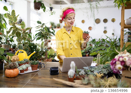 A woman in a yellow shirt holds a succulent while working on a laptop in a plant shop surrounded by greenery 136654996