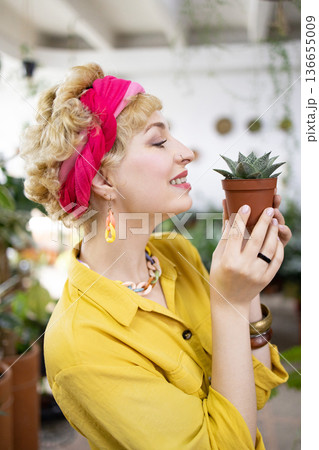 A woman smiles while holding a small potted succulent in a bright, airy greenhouse setting 136655009