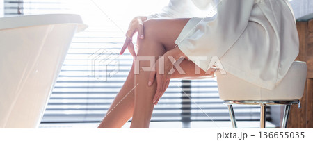 A woman sits on a stool in a bathroom. She is wearing a robe and touching her leg. Sunlight streams through blinds, creating a bright atmosphere. The space is simple with a bathtub nearby. 136655035