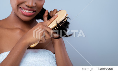 A woman is brushing her hair with a wooden brush. She is wearing a white towel and smiling. The background is light and simple, creating a relaxed scene at home. 136655061