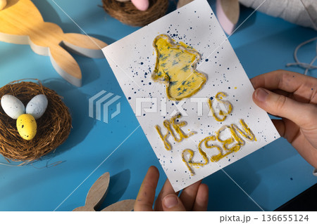 Person holding handmade greeting card with message HE IS RISEN written in beautiful handwriting. Gesture shows excitement and joy for the upcoming event. Springtime holiday gift present zero cost 136655124