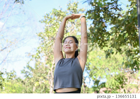 Asian woman in sportswear closing eyes standing stretching arms muscle before exercise in green park 136655131