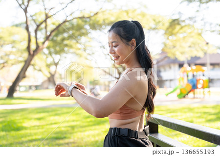 Pretty asian woman in sportswear looking at time on watch standing or resting after exercise in park 136655193