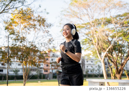 Asian woman in sportswear listening to music in headphones while running jogging exercising in park. Asian woman in sportswear listening to music in headphones while running jogging exercising in park. 136655226