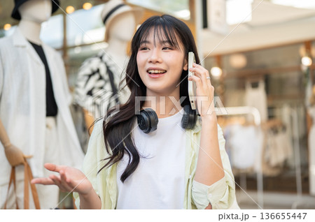 Pretty asian woman tourist talking on phone while standing or walking around the local clothing shop Pretty asian woman tourist talking on phone while standing or walking around the local clothing shop 136655447