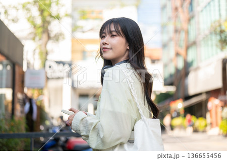 Pretty asian woman tourist holding phone carrying a bag while standing or walking around local shop. 136655456