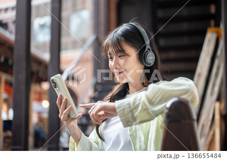 Asian woman tourist looking at phone and listening to music sitting on bench in shopping mall market Asian woman tourist looking at phone and listening to music sitting on bench in shopping mall market 136655484