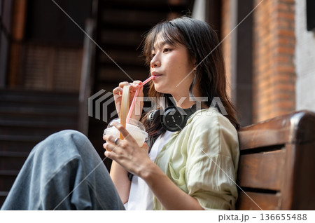 Asian woman tourist drinking fresh coconut water sitting on wooden bench in shopping mall or market. Asian woman tourist drinking fresh coconut water sitting on wooden bench in shopping mall or market. 136655488