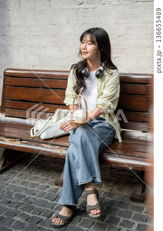 Asian woman tourist holding coconut water cup and sitting on wooden bench in shopping mall or market Asian woman tourist holding coconut water cup and sitting on wooden bench in shopping mall or market 136655489