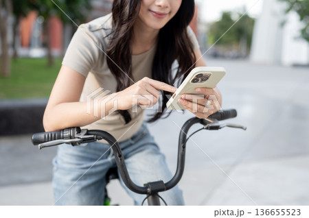 Close up of asian woman holding and looking at phone while riding on bicycle in park or city plaza. 136655523