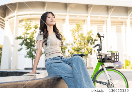 Pretty asian woman sitting and leaning back leisurely on bench aside green bicycle in park plaza. Pretty asian woman sitting and leaning back leisurely on bench aside green bicycle in park plaza. 136655529
