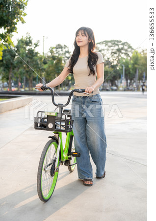 Pretty asian woman walking with green rental bicycle on pavement in park or plaza under sunlight. Pretty asian woman walking with green rental bicycle on pavement in park or plaza under sunlight. 136655533