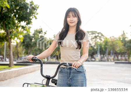 Pretty asian woman holding green rental bicycle walking on pavement in park or plaza under sunlight. 136655534